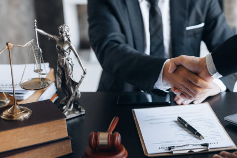 Image of a man in a suit sitting at a desk -set with a scale of justice and a statue of the Greek goddess Themis- shaking hands with a client representing how to hire a car accident lawyer.